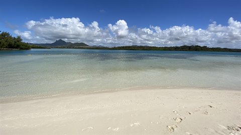 White sand before a turquoise lagoon, green mountains under white clouds, sunlight