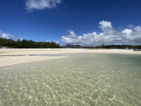 Clear shallow water over white sand, tropical beach with trees and hills under cloudy sky