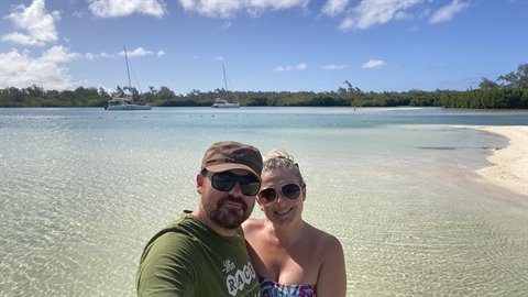 Couple takes selfie in turquoise lagoon with sailboats and pale sand