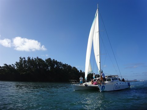White catamaran with raised sail, people on board, turquoise lagoon and tree-lined coast under blue sky