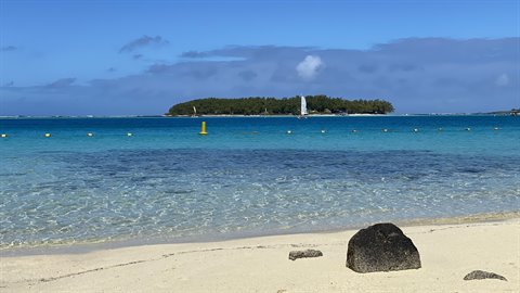 White sand and dark rocks before a green island, turquoise sea, midday light