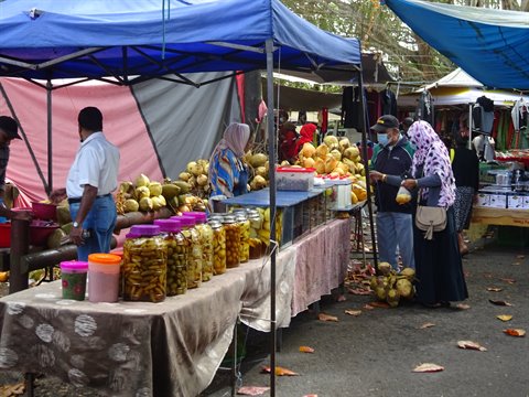 Women in headscarves browse jars and coconuts at an open-air market under blue tents