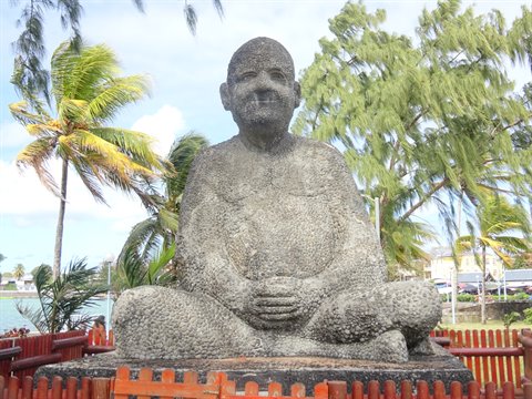 Seated stone statue in meditative pose among palm trees, daylight, red fence foreground