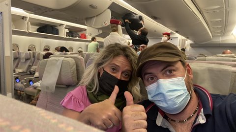 Masked couple giving thumbs up inside airplane cabin, gray seats, artificial light