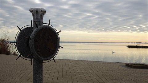 Granite column with bronze medallion on paved lakeside promenade, soft light, swan on water