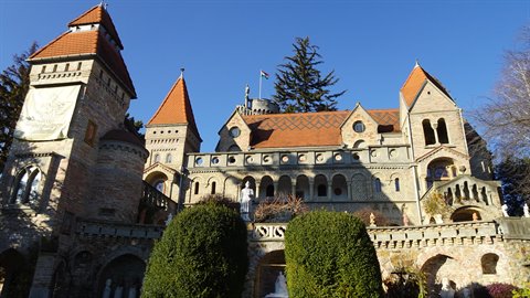 Stone medieval castle with red-roofed towers, sunlight, green shrubs in foreground