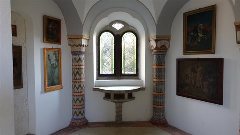 Mullioned stained-glass window between painted columns, framed paintings, natural light
