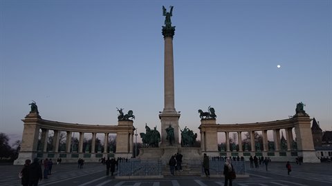 Tourists stroll beneath Budapest's millennium column in blue twilight light