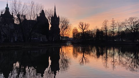 Historic castle reflected in a lake at dusk, bare trees, peach-colored sky