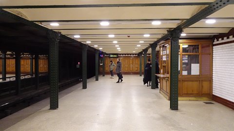 Travelers standing on a historic subway platform with green iron columns and artificial lighting