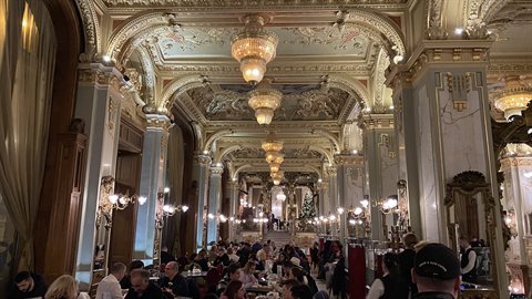 Ornate historic hall with gilded ceiling and chandeliers, visitors seated at tables under warm light