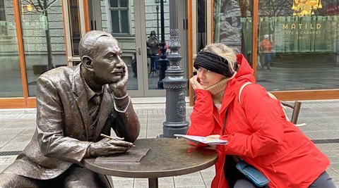 Woman in red jacket mimics a pensive bronze statue seated at a city table