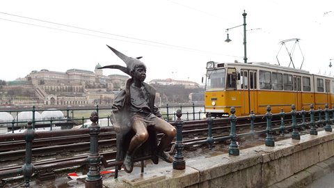 Bronze statue of a child with pointed hat on a small seat, yellow tram on tracks, soft light