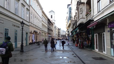 Pedestrians walking on a cobbled street lined with historic buildings and shops, church tower behind