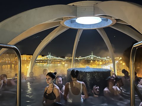 People in a rooftop thermal bath at night under lit dome, bridge over Danube behind