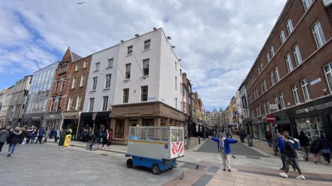Pedestrian crossing a car-free street with blue cart among brick buildings, cloudy sky