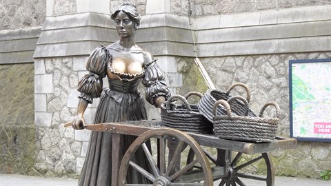 Bronze statue of a woman pushing a cart with wicker baskets beside a stone wall