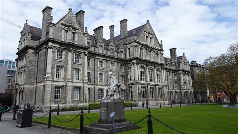 Historic grey stone college building with lawn, foreground statue, soft daylight