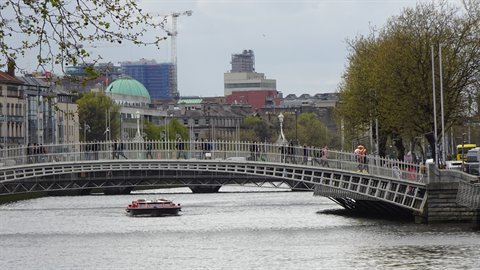 White metal footbridge over river with red tour boat, pedestrians crossing, diffused light