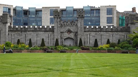 Tourist photographs grey stone castle wall before modern building, green lawn, daylight