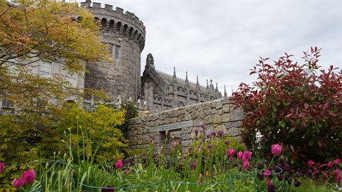 Grey stone castle turret among gardens with pink tulips under cloudy sky