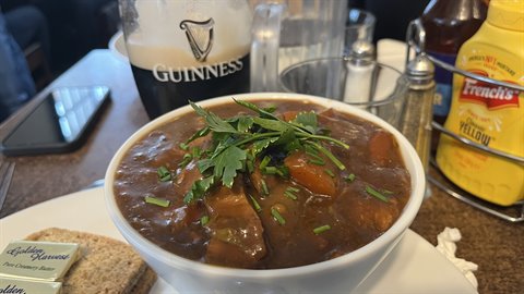 Beef stew with parsley in a white bowl, beside dark beer and bread, natural light
