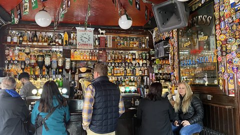 Customers at a crowded pub bar, illuminated bottles, white lamps, Guinness chalkboard