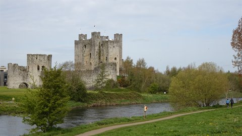 Medieval stone castle beside a river, green meadow, walkers, diffuse daylight