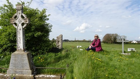 Woman sitting on green grass among stone Celtic crosses and ruins, daylight