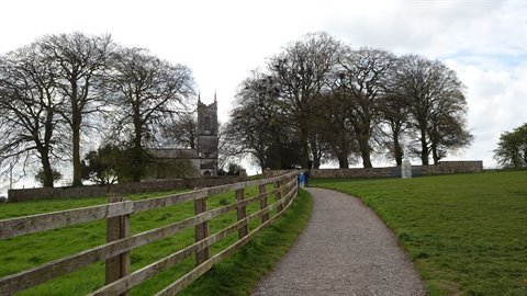 Gravel path past wooden fences leading to a stone church among bare trees, overcast light