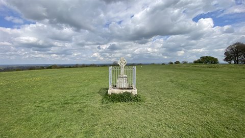 Stone Celtic cross monument enclosed by a fence in a green field, cloudy sky
