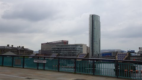 Glass skyscraper above the river, green footbridge in the foreground, cloudy sky