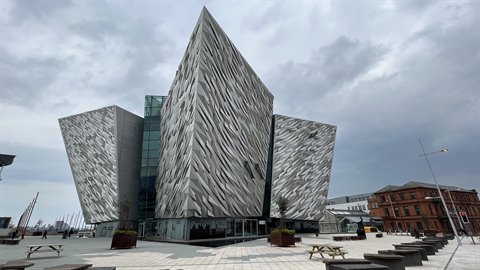 Angular museum building with grey metal facades, cloudy sky, plaza with wooden tables