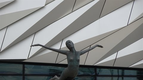Bronze female statue with outstretched arms in front of diagonal panelled facade
