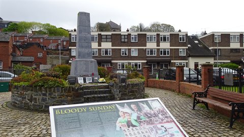 Stone memorial in a cobbled square, information panel and bench, overcast light