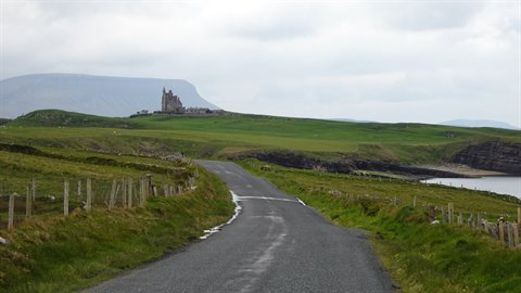 Asphalt road through green hills toward a ruined castle, diffused cloudy light