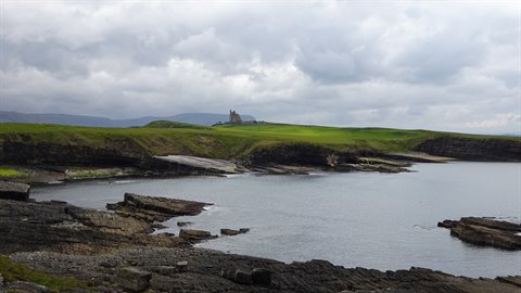 Rocky bay with calm water, green hills and an old castle under cloudy sky