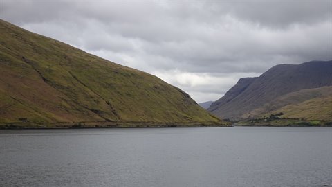 Grey lake between grassy green slopes, dark hills behind, cloudy sky