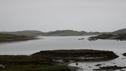 Boats resting in a still grey bay, green hills under overcast sky