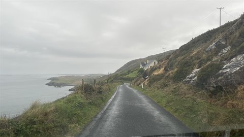 Wet paved coastal road, green hills, cloudy sky, house in the distance