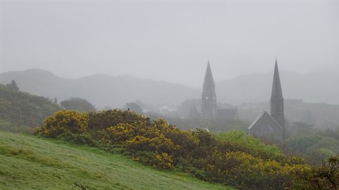 Two church spires in fog above flowering bushes and grassy hill