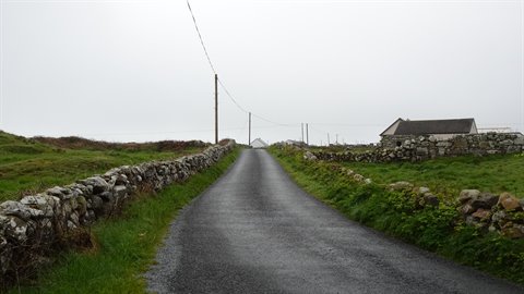 Asphalt road between dry stone walls, white houses and green fields under grey sky