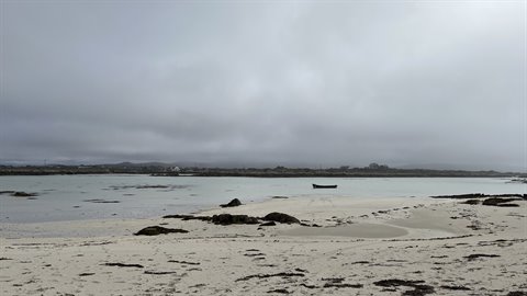 Lone boat on shallow water between sand and seaweed, grey cloudy sky