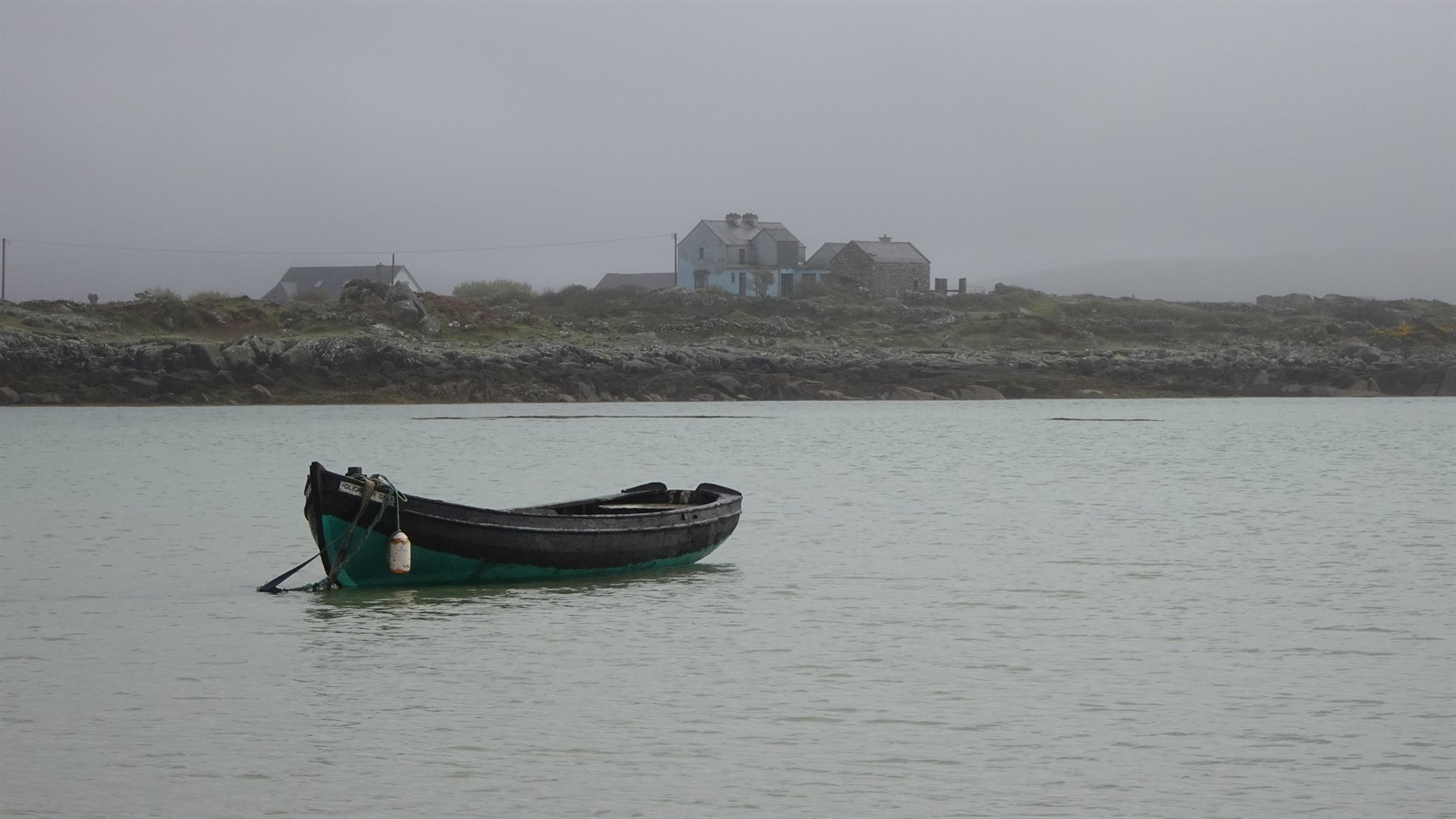 Wooden boat moored on calm water, houses on misty coast, soft light