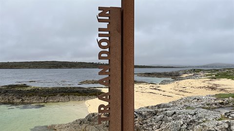Rusted vertical signpost on sandy beach with rocks, turquoise water and overcast sky