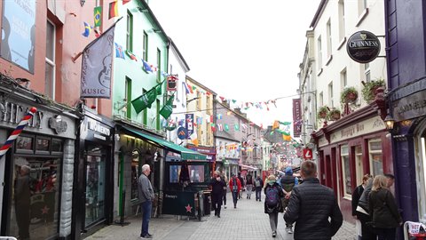 People walk along a pedestrian street lined with colourful facades, flags and pub signs