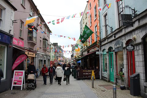Pedestrians strolling along colorful shop-lined street with hanging flags, soft daylight