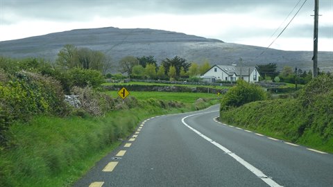 Asphalt road winding between green hedges, white house and rocky hill under cloudy sky