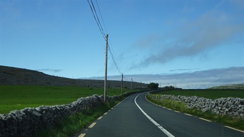 Rural asphalt road between dry stone walls and green fields, cloudy day