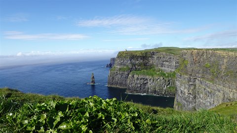 Cliffs plunging into the ocean, green grass in foreground, blue sky with clouds